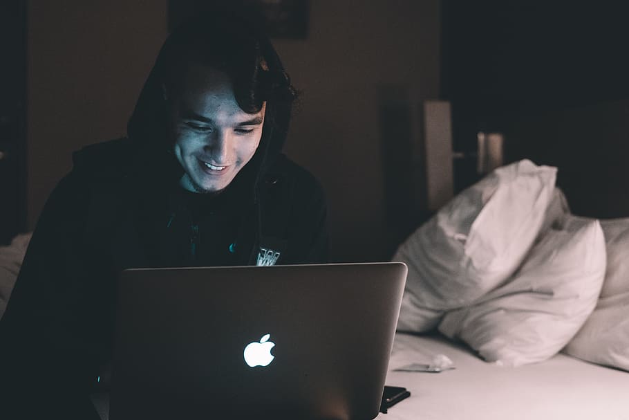Seorang pria mengenakan jaket hoodie hitam tersenyum sambil duduk di atas kasur dan membuka laptop